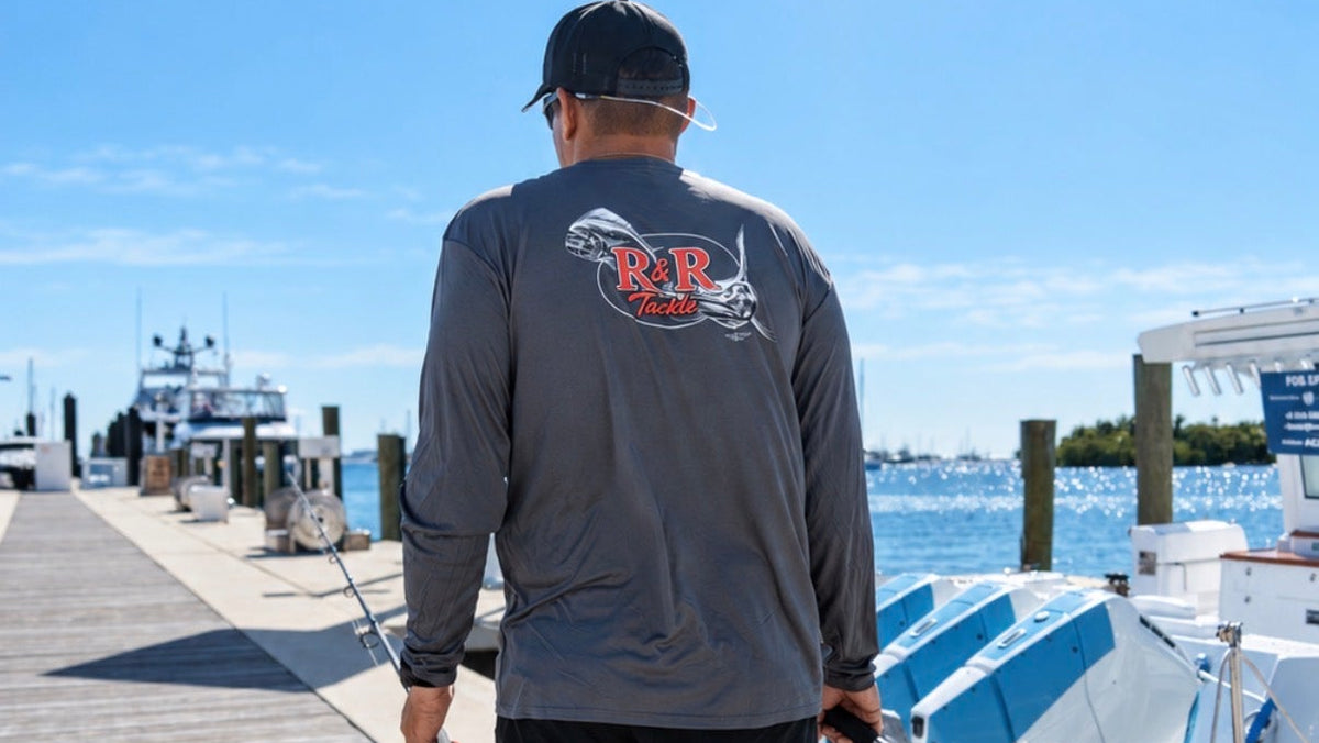 Angler wearing a gray R&R Tackle long-sleeve shirt and black hat, carrying two fishing rods along a marina dock.