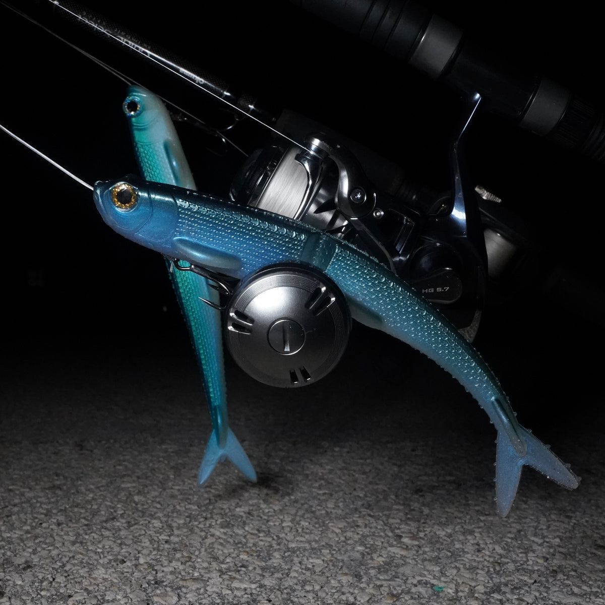 Blue R&R Tackle Naughty Lady glide baits rigged beside spinning reels, showing textured scales and forked tails.